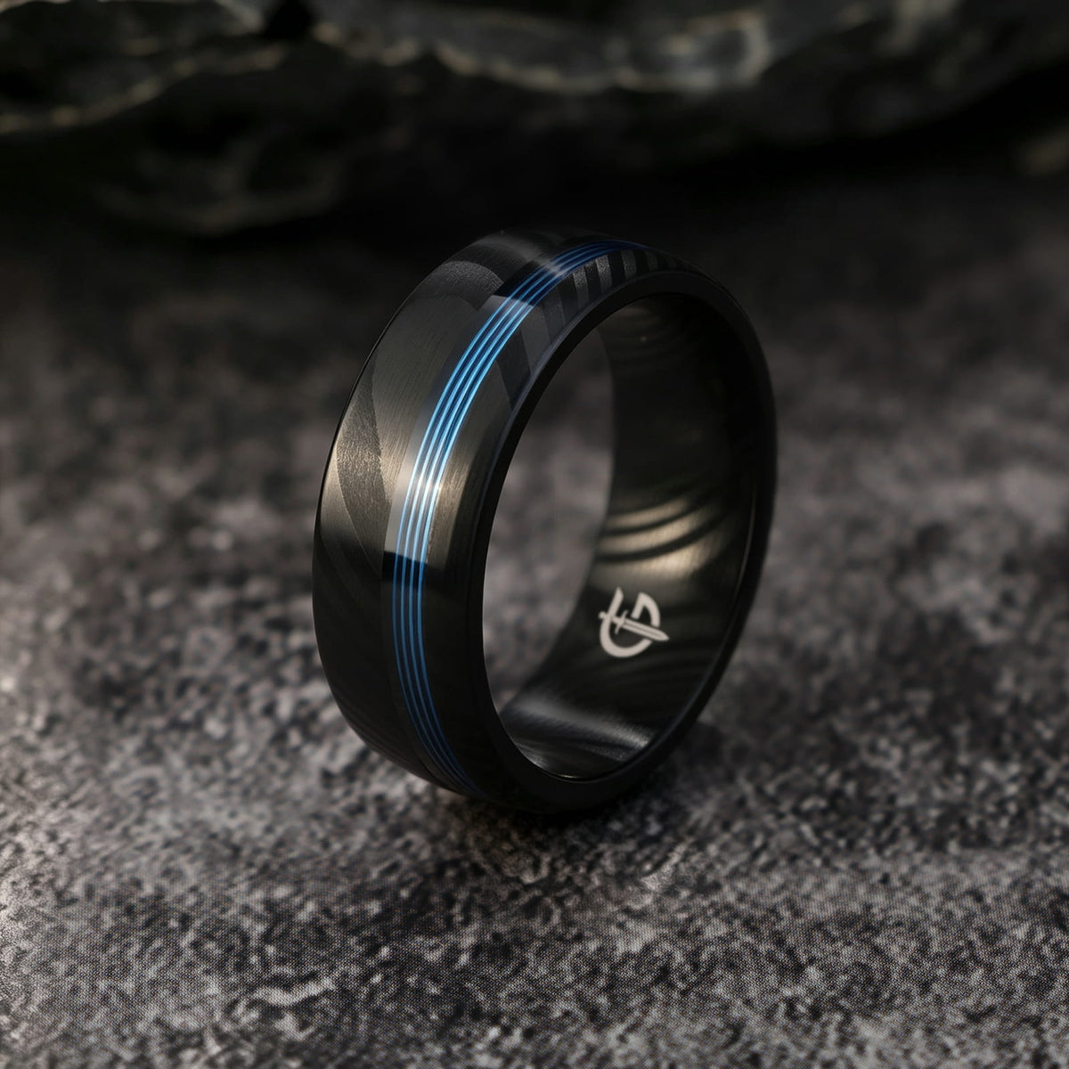 Black Damascus steel ring with diagonal gray markings and a narrow blue fishing line inlay.