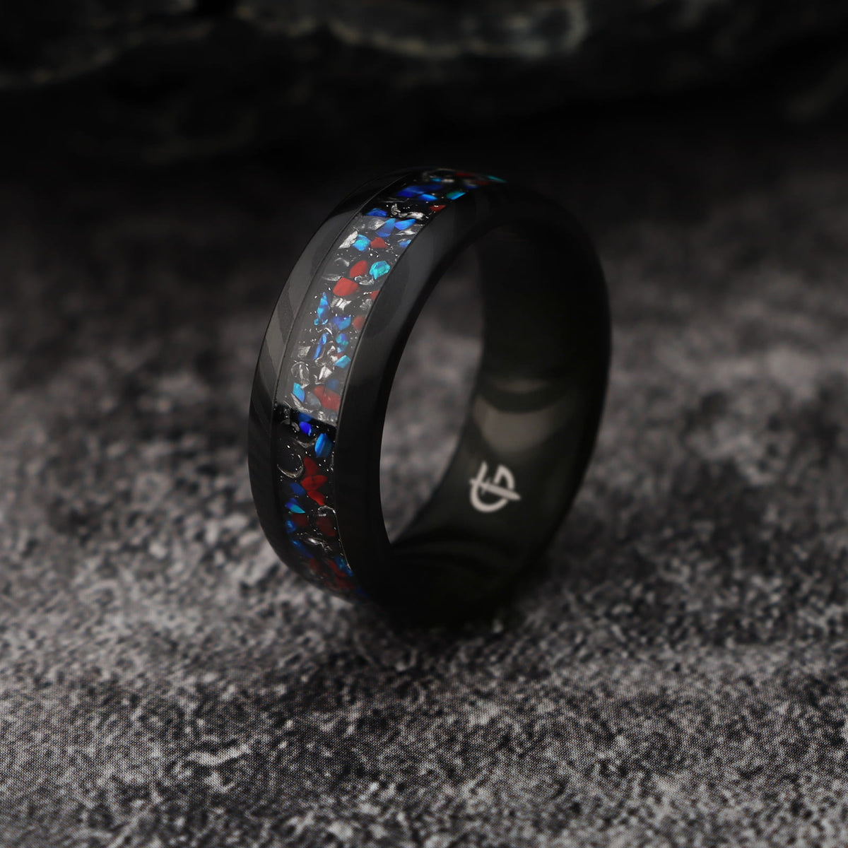 Black patterned Damascus steel ring with a central inlay of red, blue and silver-colored fragments.
