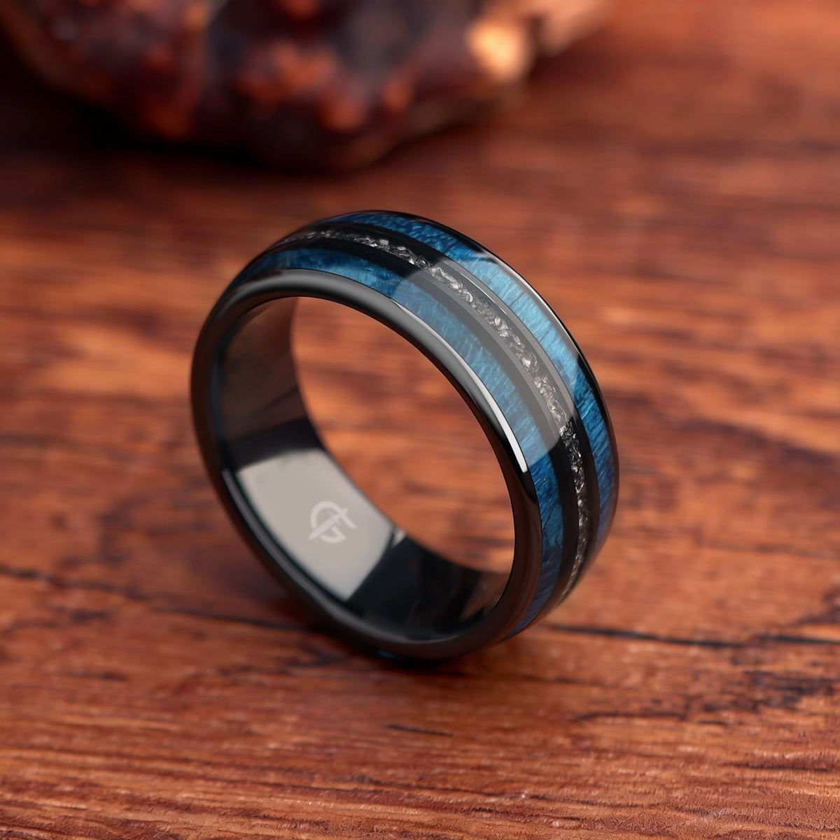 Polished black ceramic ring with two blue whiskey barrel wood bands framing silver-gray Gibeon meteorite fragments.